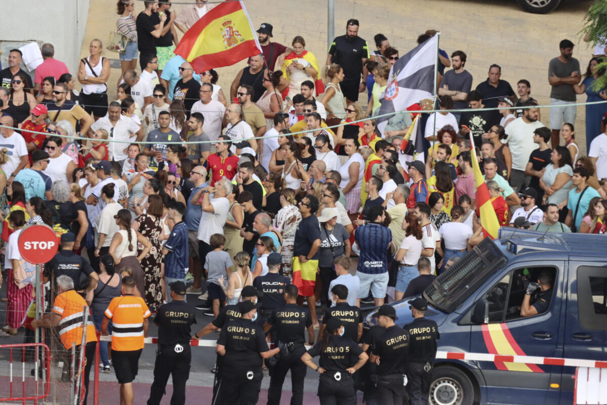 CEUTA, 27/08/2026.- Los migrantes recogen comida suministrada por voluntarios de Cruz Roja en la playa del Trampolín de Ceuta mientras la Policía Nacional impide que los manifestante lleguen al lugar donde se está llevando a cabo el reparto de las bolsas con alimentos EFE/Reduan