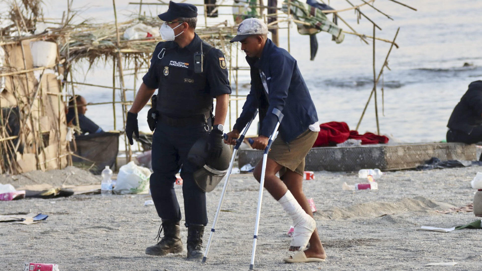 CEUTA, 20/08/2026.- Un policía acompaña a uno de los migrantes durante el desalojo de la playa del Trampolín en Ceuta este jueves. El Gobierno ha habilitado temporalmente varios espacios para la acogida de 1.800 personas migrantes que se encuentran en situación de calle en la playa del Trampolín y en la zona de Loma Colmenar, en Ceuta, que prevé ampliar progresivamente. EFE/Reduan