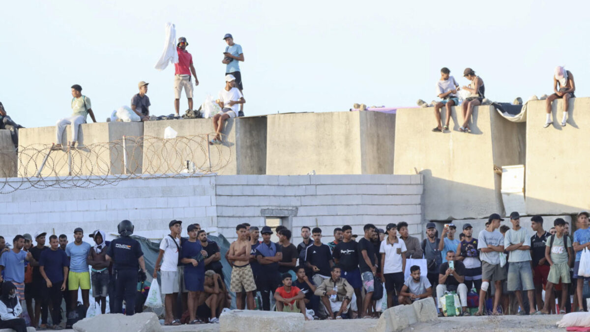 CEUTA, 27/08/2026.- Migrantes observan desde lejos las protestas de ciudadanos que impiden la llegada de los voluntarios de Cruz Roja a la playa del Trampolín con bolsas de alimentos. EFE/Reduan