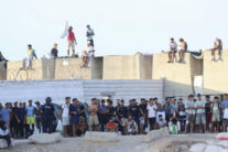 CEUTA, 27/08/2026.- Migrantes observan desde lejos las protestas de ciudadanos que impiden la llegada de los voluntarios de Cruz Roja a la playa del Trampolín con bolsas de alimentos. EFE/Reduan