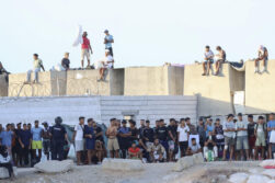CEUTA, 27/08/2026.- Migrantes observan desde lejos las protestas de ciudadanos que impiden la llegada de los voluntarios de Cruz Roja a la playa del Trampolín con bolsas de alimentos. EFE/Reduan