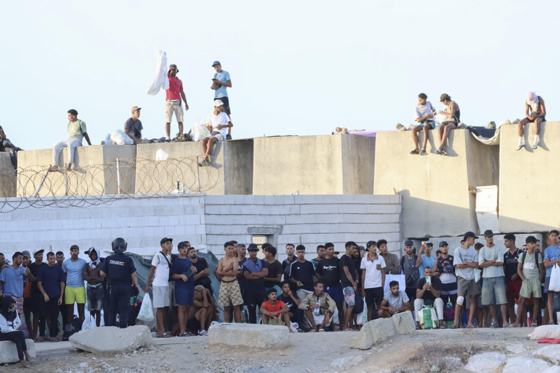CEUTA, 27/08/2026.- Migrantes observan desde lejos las protestas de ciudadanos que impiden la llegada de los voluntarios de Cruz Roja a la playa del Trampolín con bolsas de alimentos. EFE/Reduan