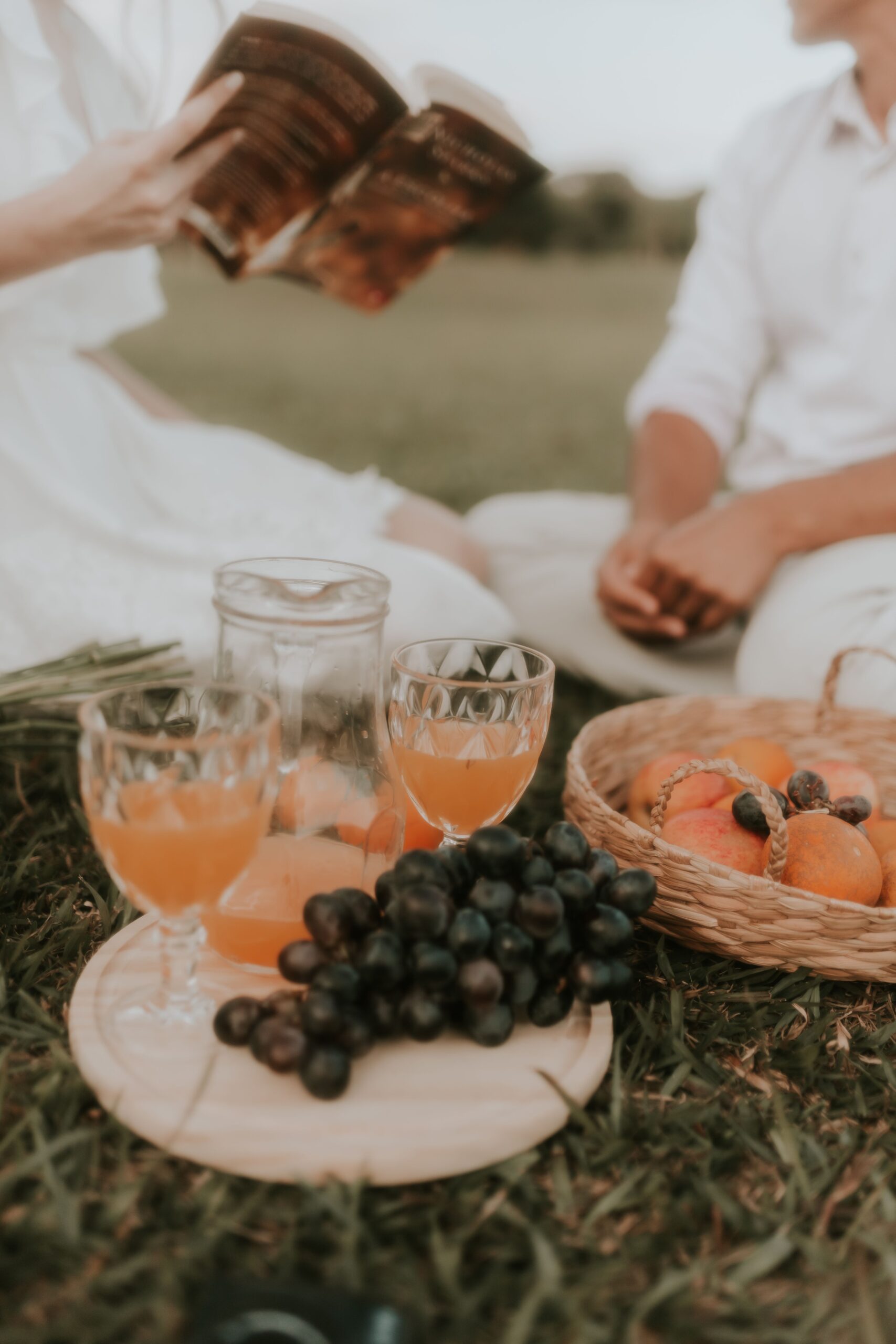A couple enjoys a picnic with fruit, drinks, and a book in a summer park setting.