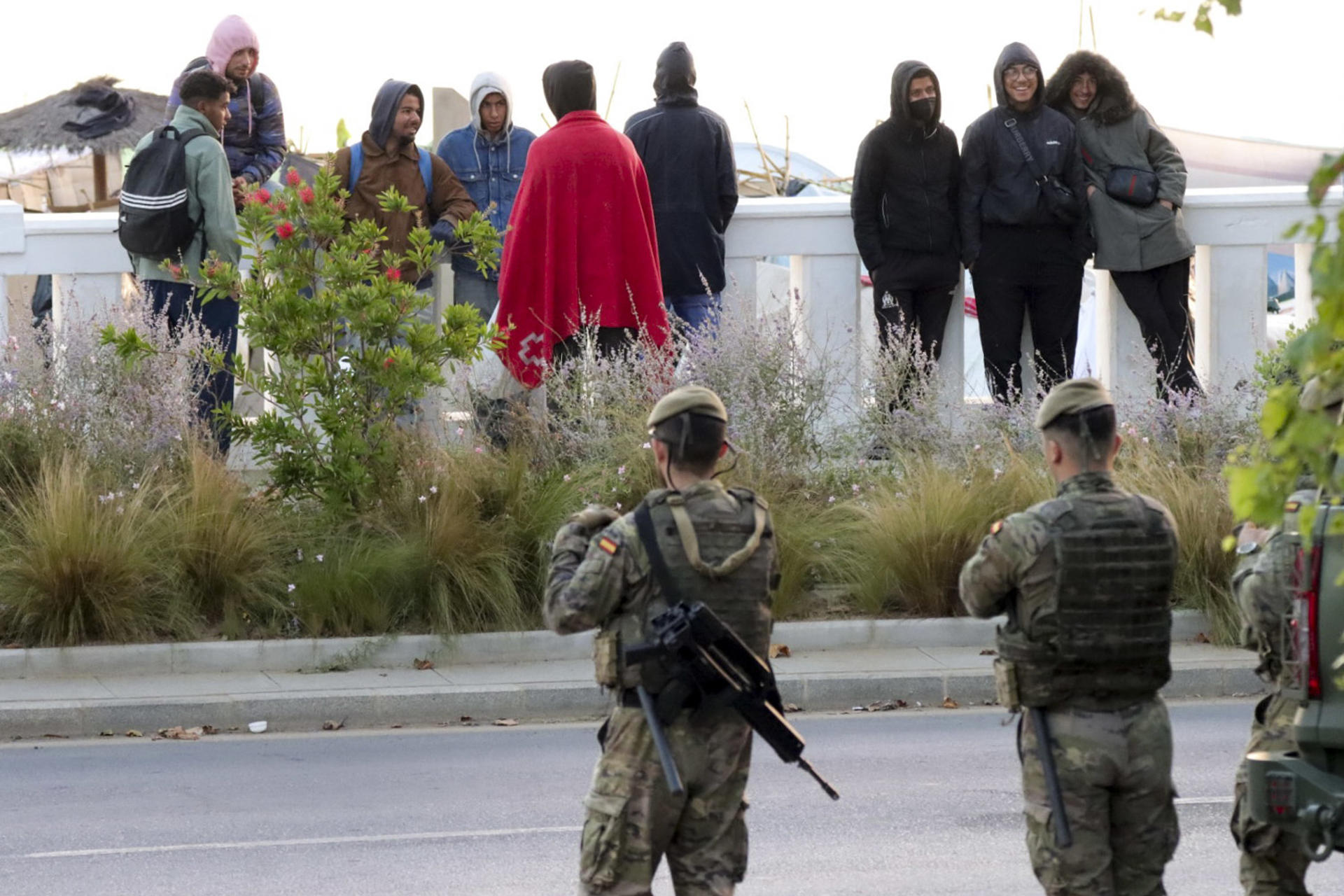 CEUTA (ESPAÑA), 16/09/2026.- Miembros del ejército observan a unos migrantes que viven en las chozas de la playa Benítez, a la espera de ser en el nuevo campamento temporal del muelle de Poniente del puerto de Ceuta para migrantes que previsiblemente se inaugura este miércoles. EFE/Reduan