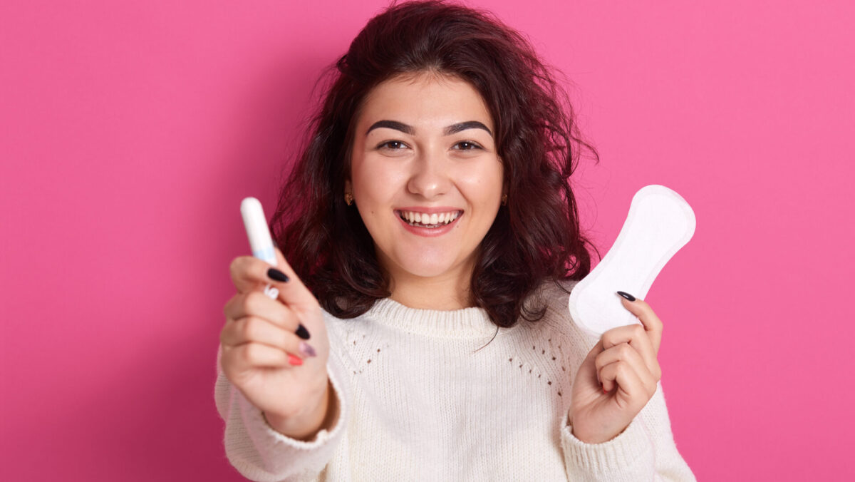 Happy satisfied Caucasian woman holding different types of feminine hygiene products during menstrual cycle, sanitary pad and cotton tampon in fwemales hands, standing isolated over pink background.