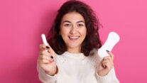 Happy satisfied Caucasian woman holding different types of feminine hygiene products during menstrual cycle, sanitary pad and cotton tampon in fwemales hands, standing isolated over pink background.