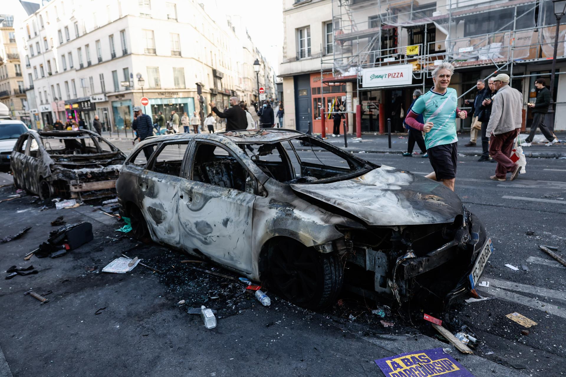 Paris (France), 18/10/2026.- People walk past a burnt out car in the aftermath of clashes between riot police and demonstrators in Paris, France, during nationwide protests against the high cost of living in the country, 18 October 2026. (Protestas, Francia) EFE/EPA/MOHAMMED BADRA
